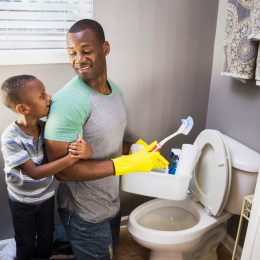 A young boy hugging the arm of a man cleaning a toilet while wearing yellow gloves and holding cleaning supplies
