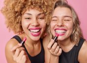 Close up of two friends smiling and putting on pink lipstick.