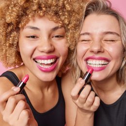 Close up of two friends smiling and putting on pink lipstick.