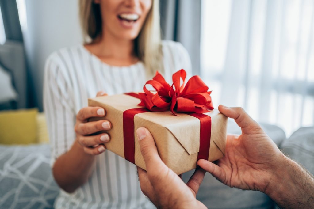 Cropped shot of a loving husband giving his wife a gift. Boyfriend surprise his beautiful girlfriend with present while she is sitting on the sofa in the living room at home. Focus is on the gift.