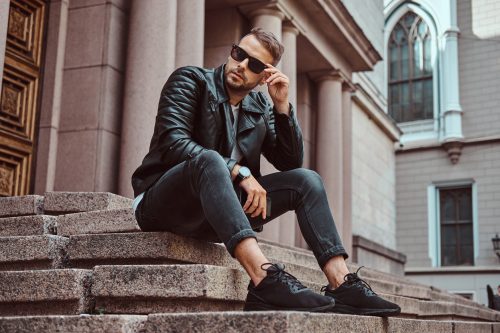 Fashionable guy dressed in a black jacket and jeans holds a smartphone sitting on steps against an old building in Europe.