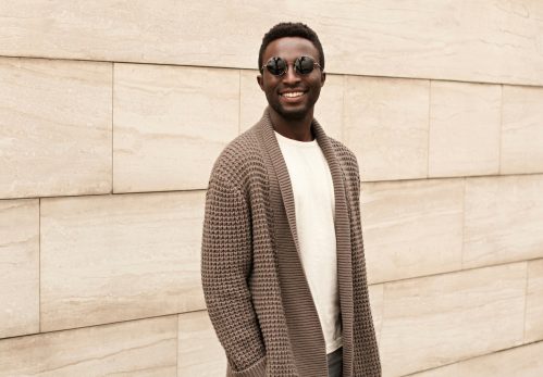 Stylish smiling african man wearing brown knitted cardigan and sunglasses on city street over brick wall background