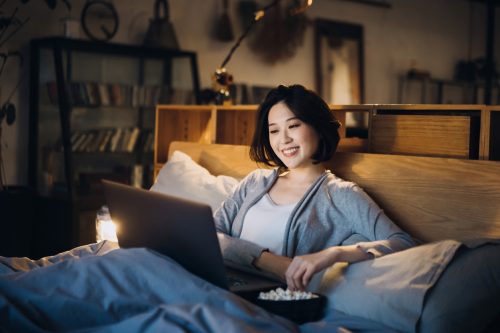 Cheerful young Asian woman lying on bed enjoying the weekend, watching movie on laptop and eating popcorn at home in the evening