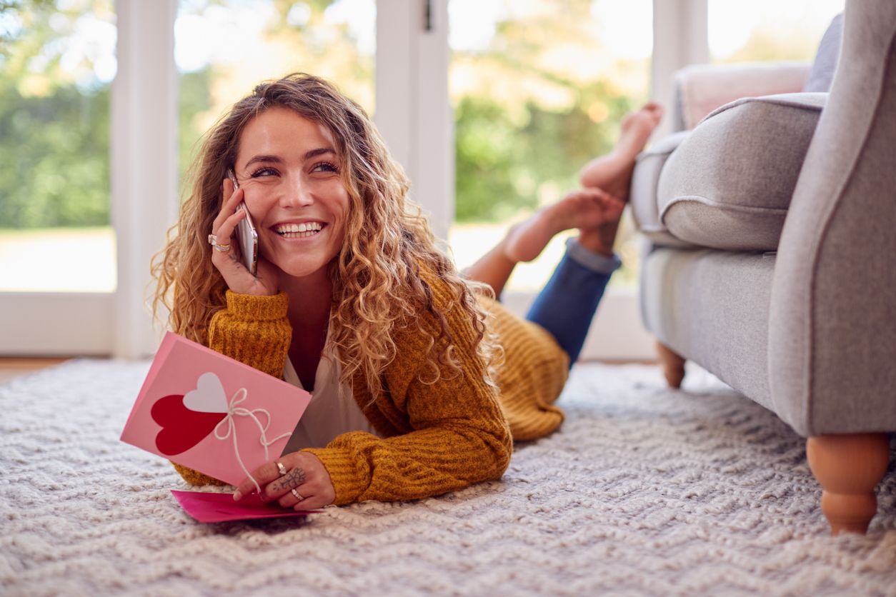 Woman Lying On Floor At Home Reading Valentine's Day Card And Talking On Mobile Phone