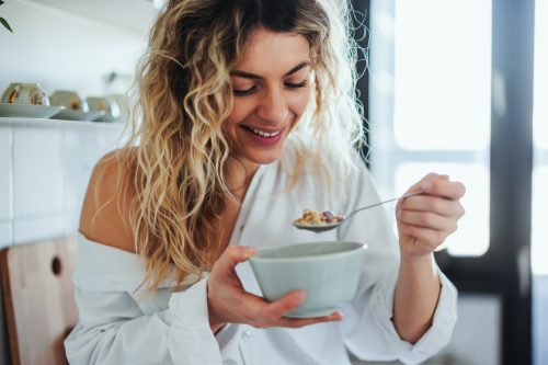 Young woman in pajamas is eating oatmeal for breakfast in her apartments kitchen