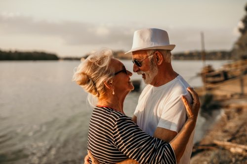 Spontaneous image of a senior couple, enjoying their sunset beach walk together. A cool looking grandpa, wearing a hat, is hugging his beautiful wife, both wearing sunglasses on, smiling and facing each other, feeling romantic, dancing. Enjoying conversations, love, and small dance
