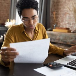 Focused young woman in eyeglasses looking through paper documents, managing business affairs, summarizing taxes, planning future investments, accounting alone at home office.