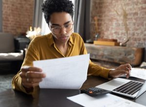 Focused young woman in eyeglasses looking through paper documents, managing business affairs, summarizing taxes, planning future investments, accounting alone at home office.