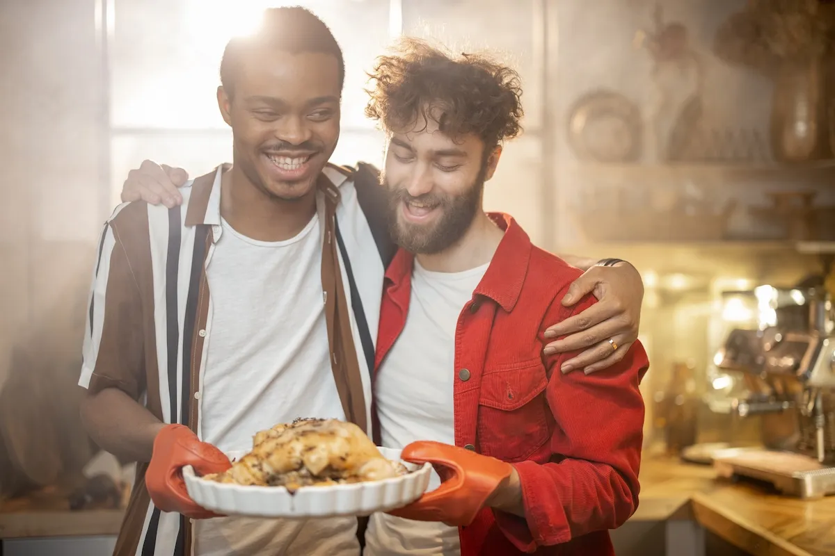 A male couple hugging and holding a dish of chicken in their kitchen.