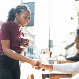 A man sitting at an outdoor restaurant table shakes hands with a woman who he's meeting for a first date.