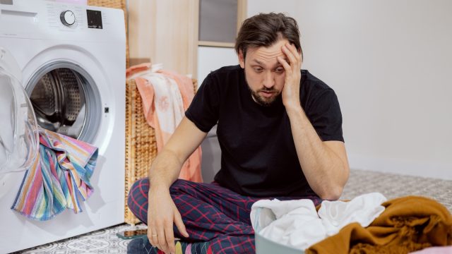 Man sits on the laundry room floor with the washing machine open, looking confused and overwhelmed.