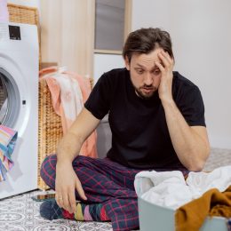 Man sits on the laundry room floor with the washing machine open, looking confused and overwhelmed.