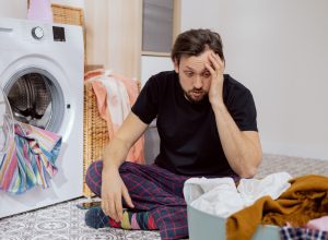 Man sits on the laundry room floor with the washing machine open, looking confused and overwhelmed.