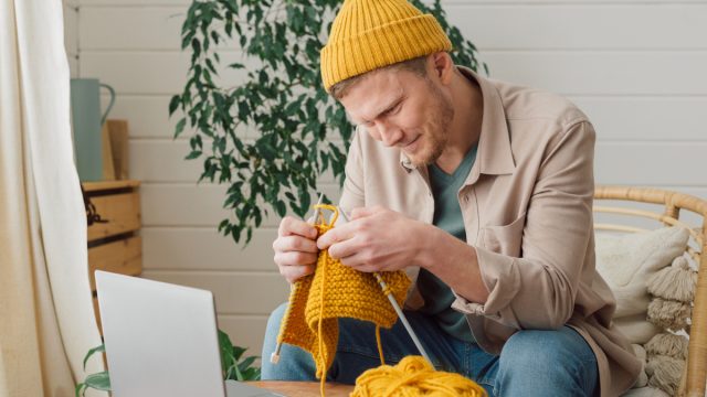 A man watching a video to learn how to knit a yellow scarf to match his yellow hat.