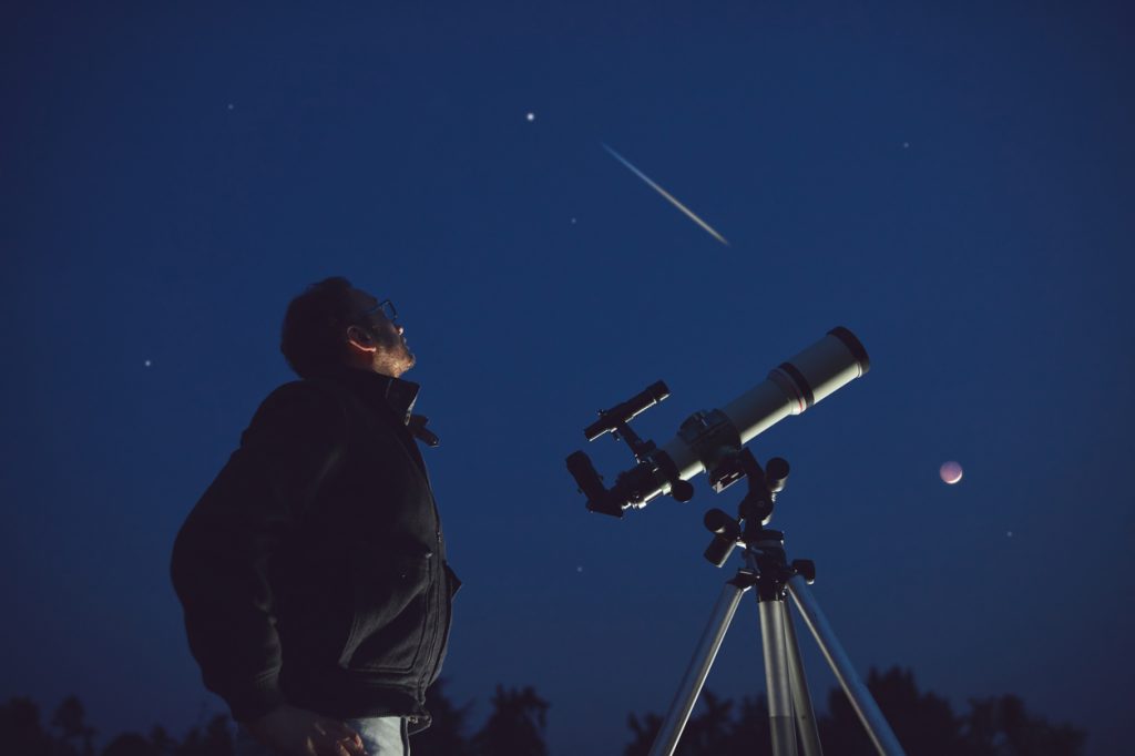 The silhouette of a person looking up at the night sky with a telescope