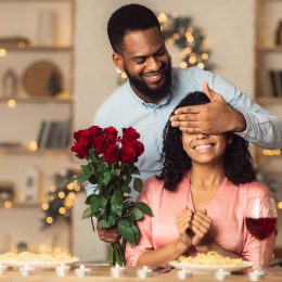 A man covering a woman's eyes at the dinner table so he can surprise her with red roses.