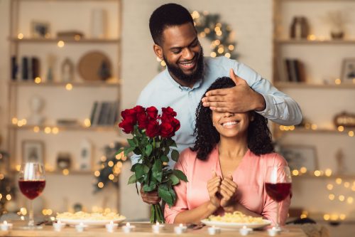 A man covering a woman's eyes at the dinner table so he can surprise her with red roses.