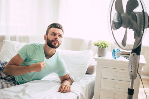 Sweaty young man is trying to refresh from the heat with a fan while lying in bed at home.