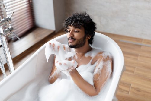 Handsome indian man blowing soap bubbles while relaxing in bathtub with foam
