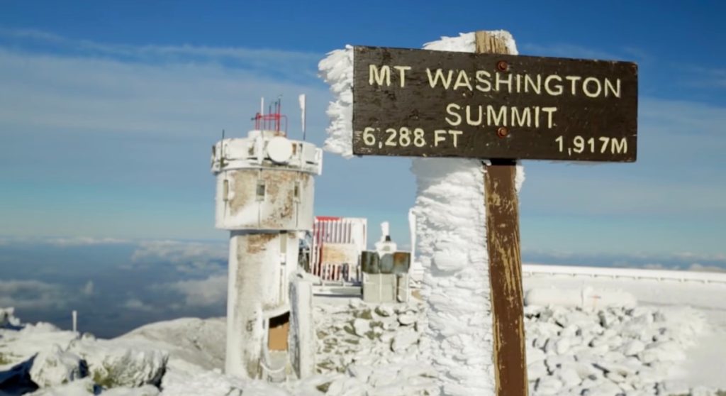 The Mount Washington Observatory summit with a sign covered in snow that reads an elevation of 6,220 feet