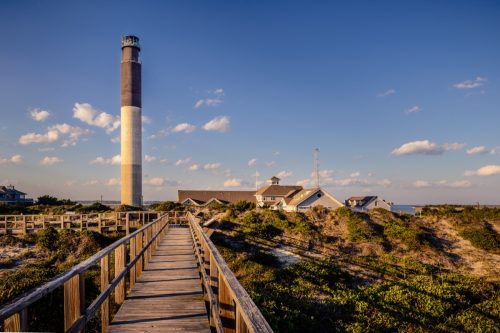 A walking ramp that leads the view to a majestic lighthouse in Oak Island Beach in North Carolina. This image was taken during a sunny afternoon in spring season.