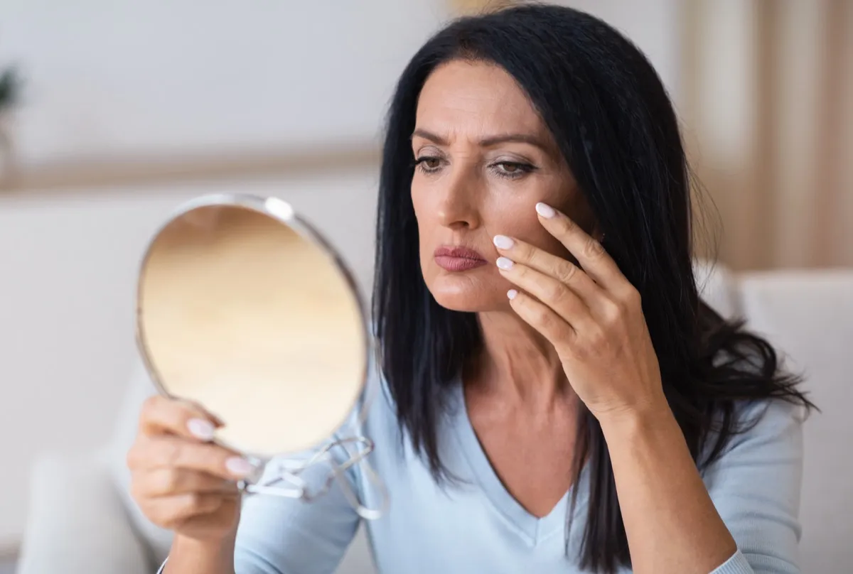 Portrait of serious senior woman with long dark black hair holding mirror and looking at her reflection, cheking and examining facial wrinkles, doing morning skincare routine at home