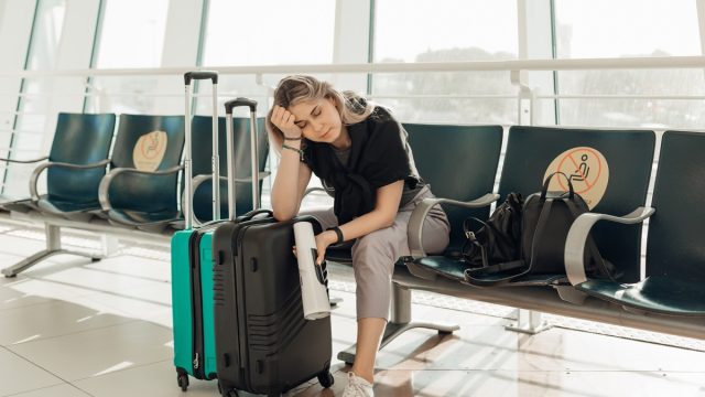 Bored blond woman with luggage, leaning elbow on bags, sitting in waiting room at airport due to coronavirus pandemic Covid-19 outbreak travel restrictions. Flight cancellation. Too late for voyage