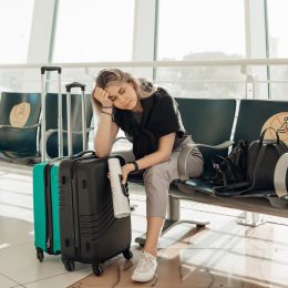Bored blond woman with luggage, leaning elbow on bags, sitting in waiting room at airport due to coronavirus pandemic Covid-19 outbreak travel restrictions. Flight cancellation. Too late for voyage