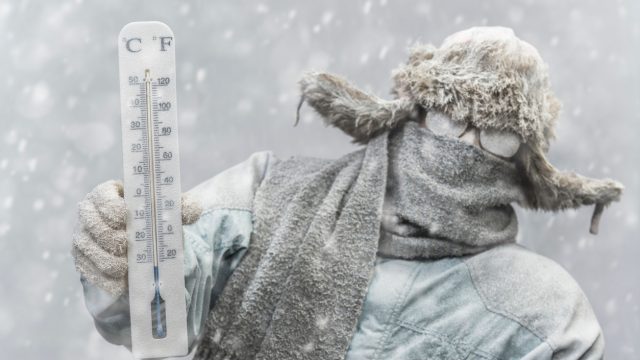 A person holding a thermometer in freezing weather while bundled from head to toe and covered in frost