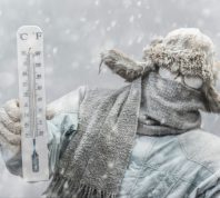 A person holding a thermometer in freezing weather while bundled from head to toe and covered in frost