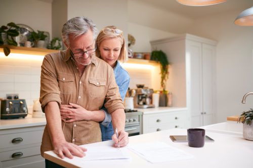 An older middle-aged couple standing in their kitchen and looking over investment documents while hugging