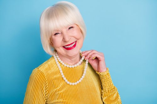 A senior woman with white hair wearing a yellow shirt, pearl necklace, and red lipstick against a blue background