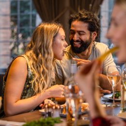group of friends wearing smart-casual clothing in a restaurant in the city on a winter's day. They are all sitting around a table and enjoying food and drinks. One of the men is feeding his partner some of his food.