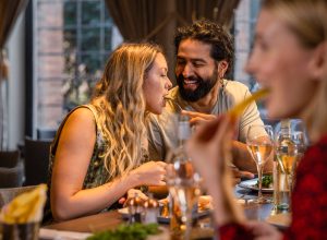 group of friends wearing smart-casual clothing in a restaurant in the city on a winter's day. They are all sitting around a table and enjoying food and drinks. One of the men is feeding his partner some of his food.