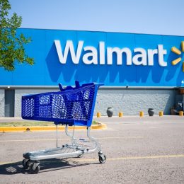Shopping cart on a parking lot in front of main entrance to Walmart supermarket outdoor on the street with no people. Big Walmart logo on blue background behind.