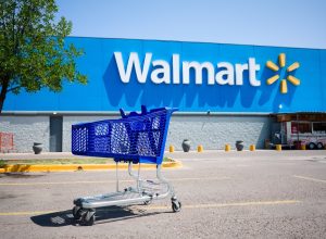 Shopping cart on a parking lot in front of main entrance to Walmart supermarket outdoor on the street with no people. Big Walmart logo on blue background behind.