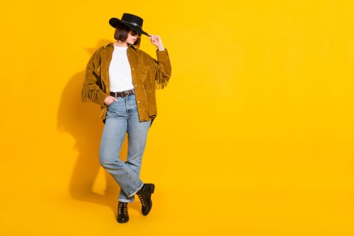 Young woman wearing a black cowboy hat, a brown suede fringe jacket, jeans, and black boots—against a yellow background.