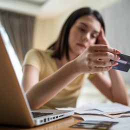 A young woman holding a credit card at her desk and looking at it with a sad expression