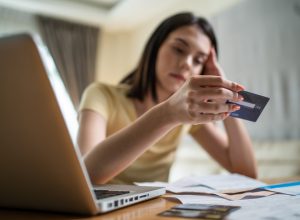 A young woman holding a credit card at her desk and looking at it with a sad expression