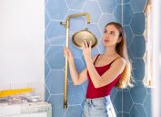 A young woman putting up a brass shower head in an under-construction bathroom.