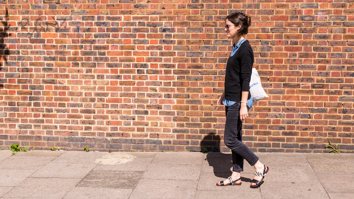 Young trendy woman wearing sandals, black skinny jeans, blue blouse, black sweater and sunglasses walking on a street in springtime. Red brick wall as background.