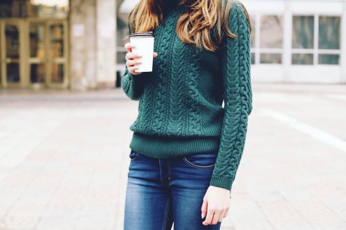 View from the neck down of a woman with long brown hair wearing a green cable knit sweater and jeans, holding a coffee cup.