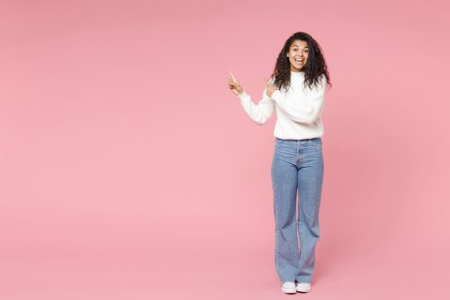 A smiling woman wearing a white turtleneck sweater, jeans, and white converse sneakers against a pink background