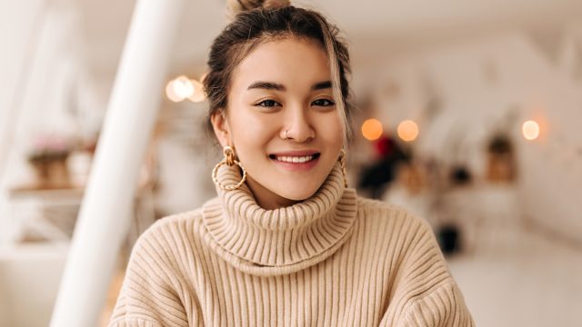 A smiling young woman wearing a beige turtleneck sweater with gold hoop earrings and her hair up in a bun.