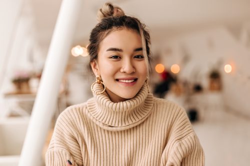 A smiling young woman wearing a beige turtleneck sweater with gold hoop earrings and her hair up in a bun.