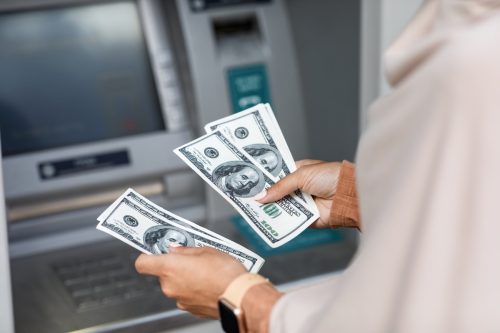 Hands of unrecognizable young business woman counts dollars near atm with empty screen, cropped. Online payment, money transfers, financial transactions and digital financial services