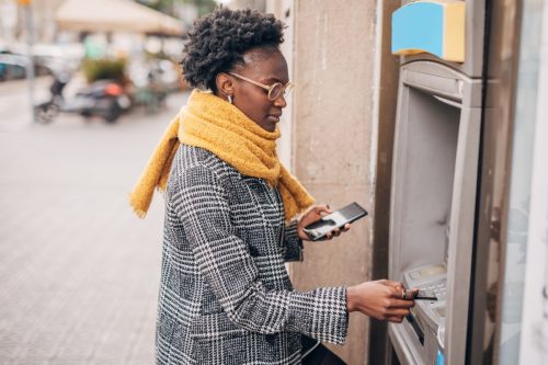 Young woman talking on the mobile phone and using ATM and taking cash from the card