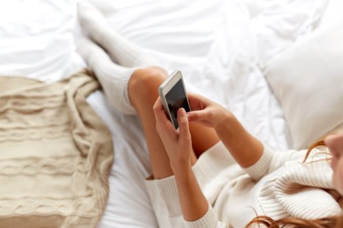 Close up on legs and hands of woman reading her phone in bed