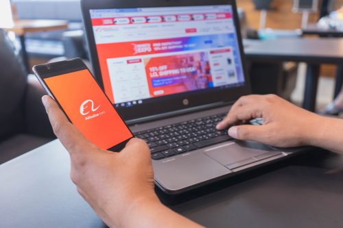A person holds a phone with the Alibaba app open while shopping at Alibaba on their laptop.