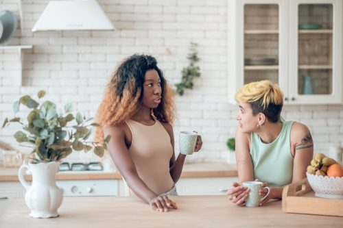 Two young adult concerned girlfriends in casual clothes communicating drinking coffee in kitchen at home